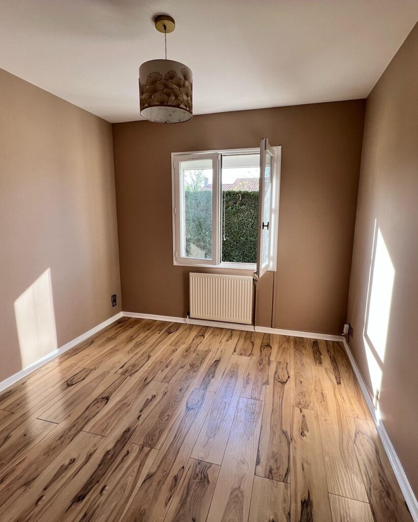 Chambre moderne : Parquet bois clair, murs taupe. Chambre rénovée murs taupe, parquet bois clair contrasté. Lumière solaire chaude, fenêtre ouverte. Luminaire ginkgo doré au plafond.