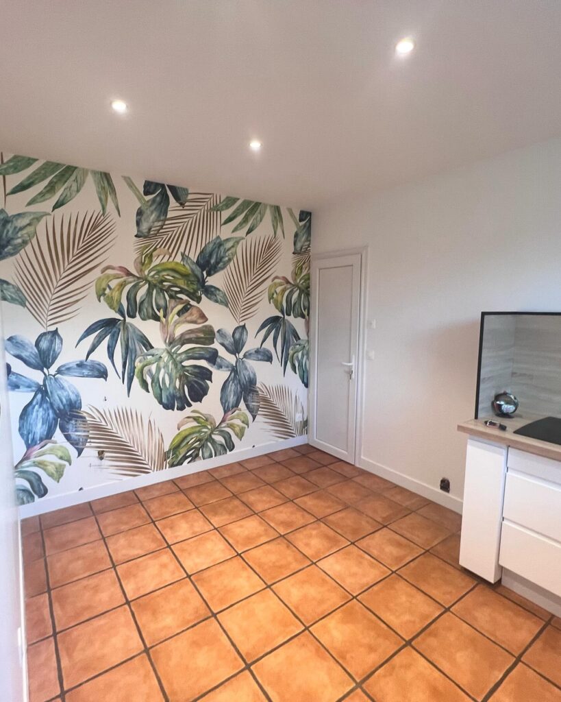 Papier peint tropical chic, carrelage terracotta et intérieur moderne. Intérieur moderne avec mur d'accent tropical (feuilles vertes et bleues) et porte blanche. Le sol est recouvert de grands carreaux de terracotta.