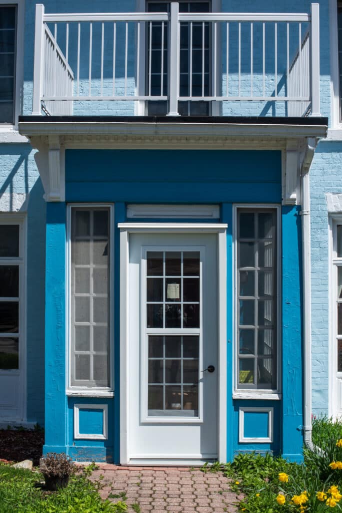 Façade bleue vive : Entrée avec porte blanche et balcon Entrée de maison bleu vif avec porte blanche à carreaux, entourée de fenêtres et surplombée par un balcon blanc. Note visible dans la vitre.